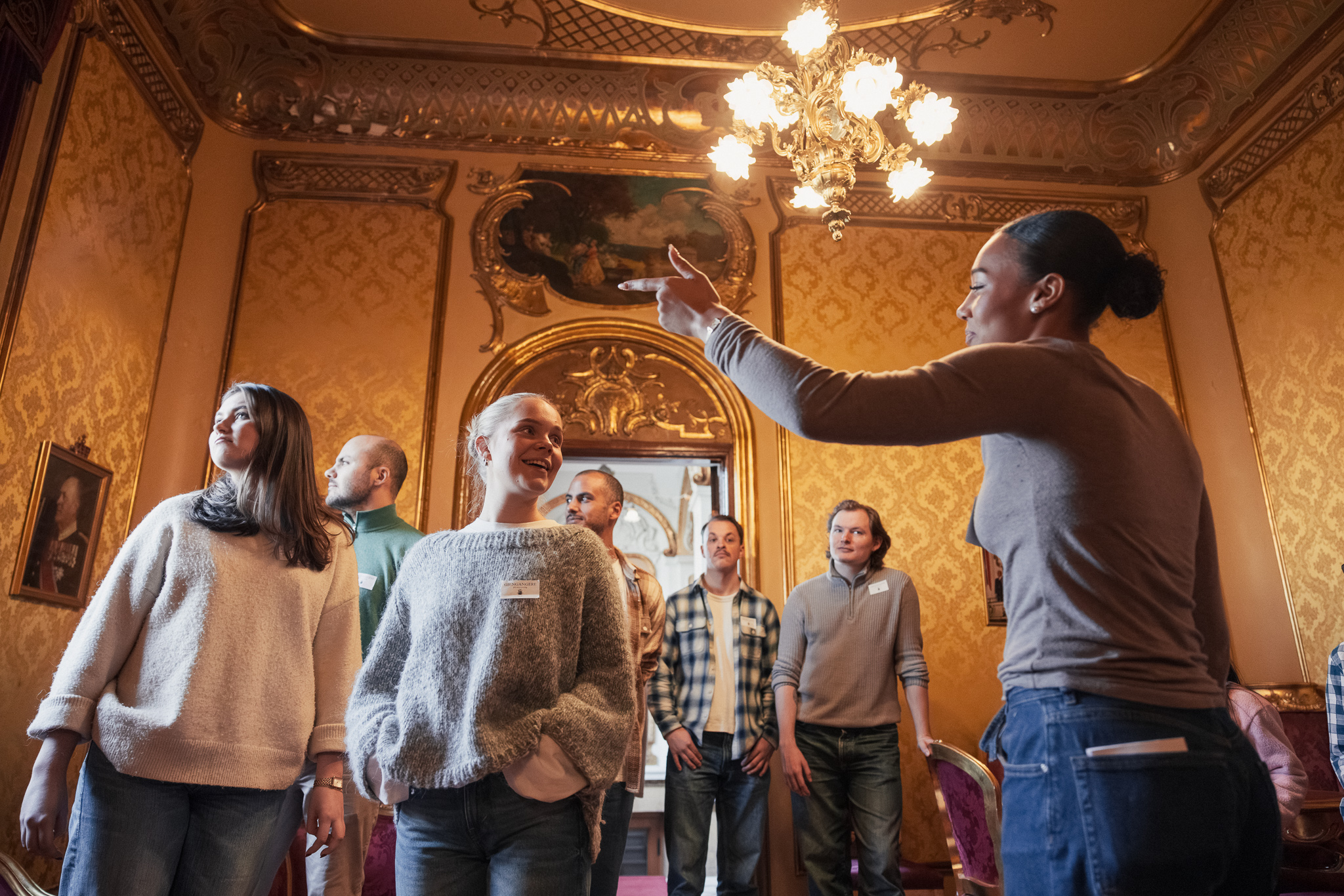 Omviser gestikulerer og snakker til en gruppe voksne i et rom med gyldne vegger og lysekrone.